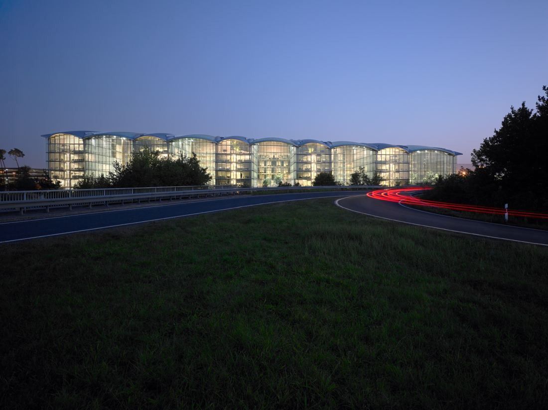Lufthansa Group: Glass building with arched roofs glowing at dusk behind a grassy embankment and road