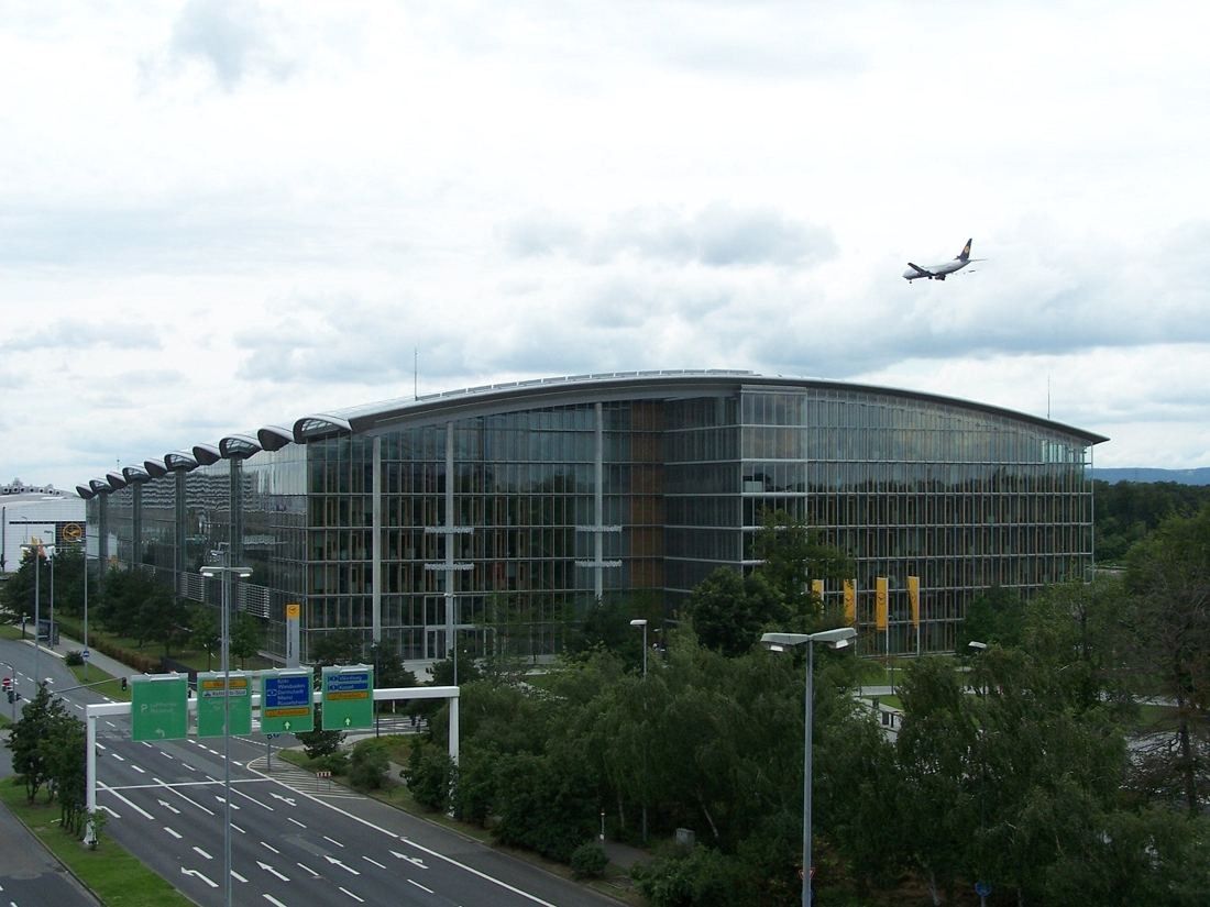 Lufthansa Group: Long glass building with a curved roof beside a highway, an airliner flying overhead