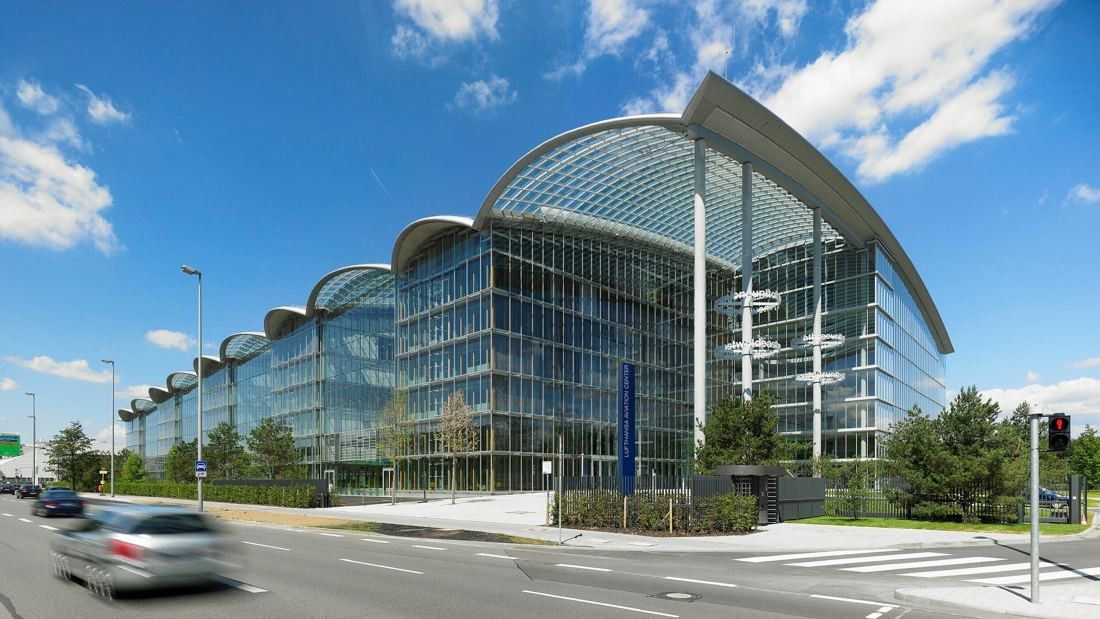 Long glass office building with curved roof arches beside a road on a sunny day