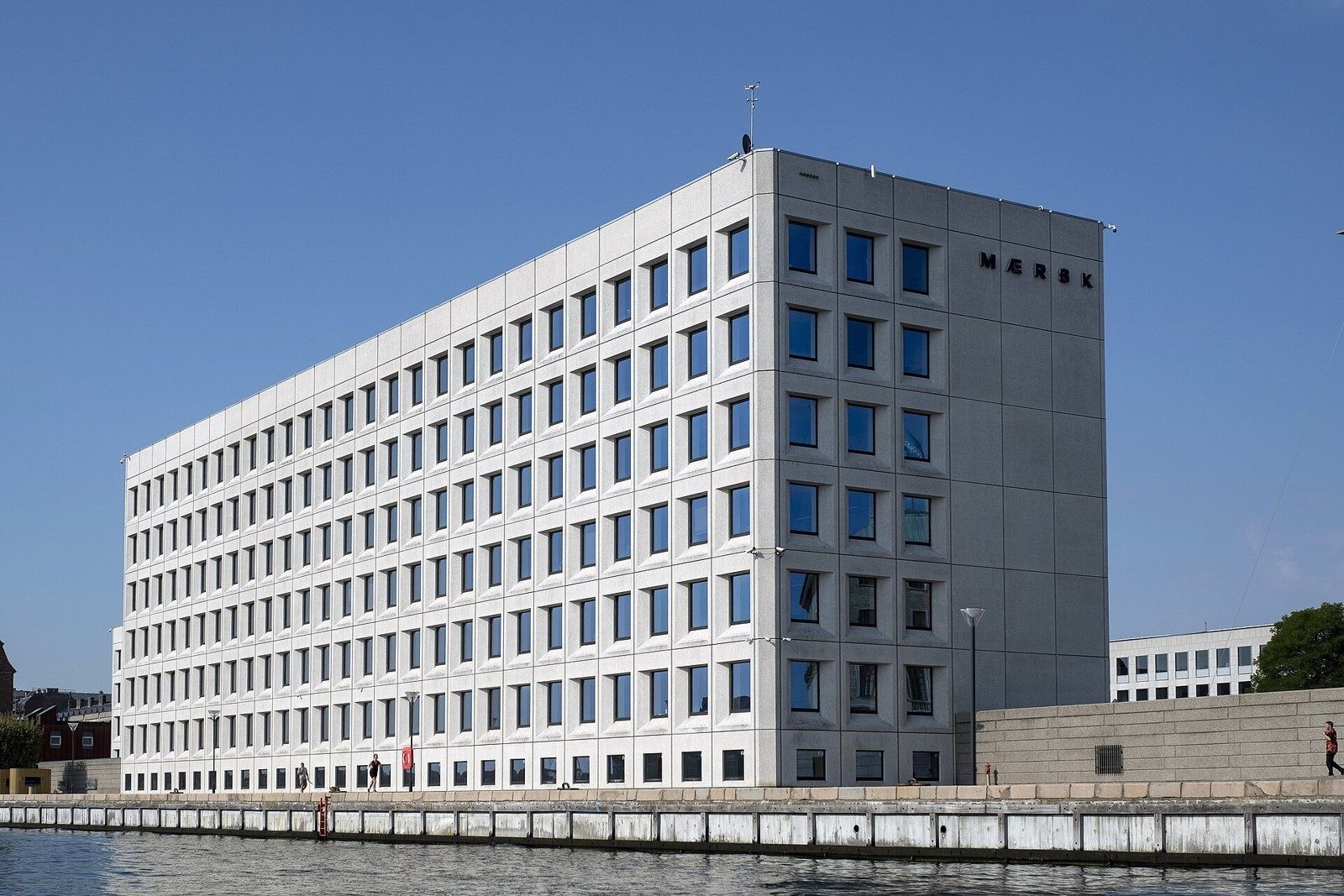 Large pale concrete office block with a grid of square windows on the harbour edge under a blue sky