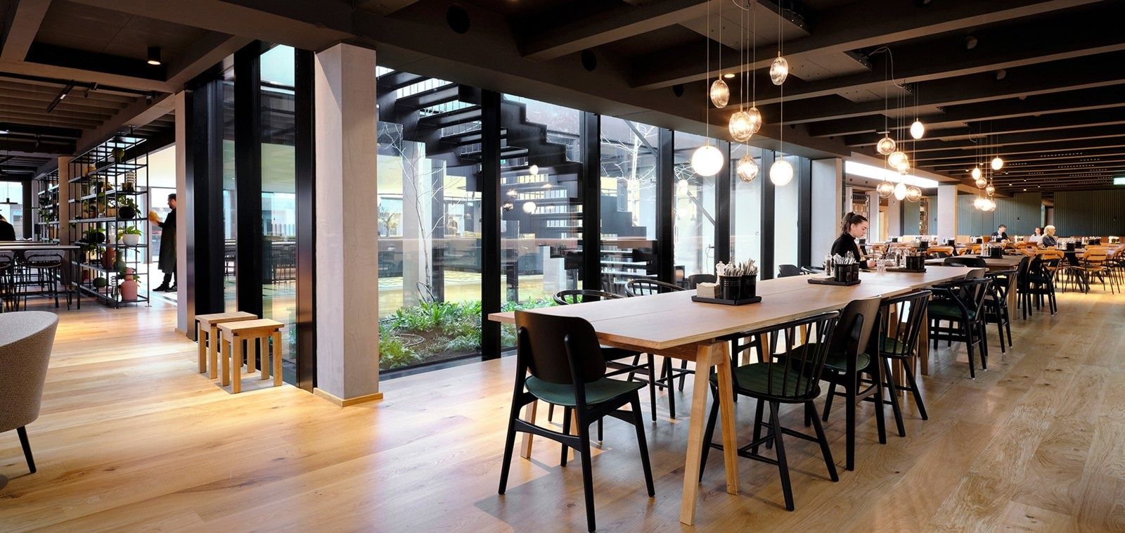 A.P. Moller - Maersk: Long communal tables in a dining hall with glass walls onto a planted courtyard and a black staircase