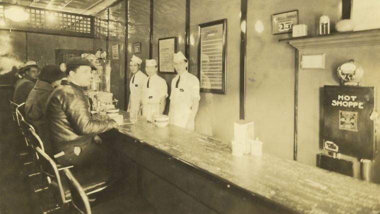 Sepia photo of a small diner counter with stools and staff behind it
