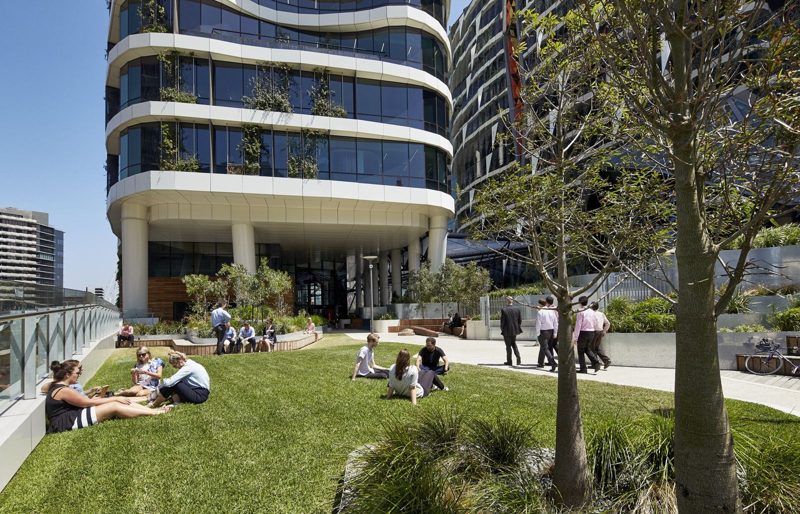 Medibank: People sitting on a sunny lawn at the foot of a curving glass tower with planted balconies