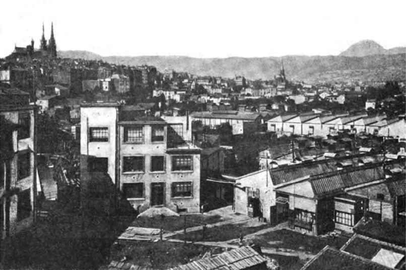 Old black-and-white view over factory roofs and multi-storey works buildings with the city behind