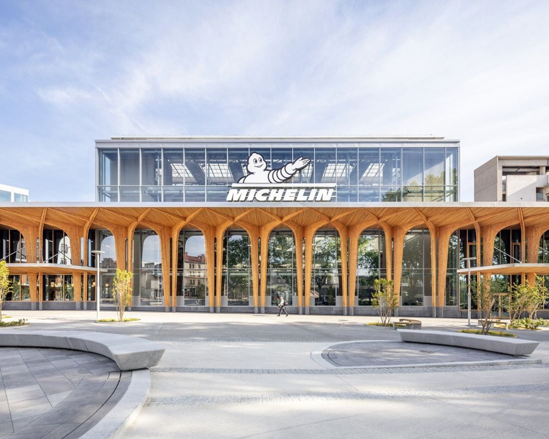 Michelin: Timber-arched façade with a glass upper storey carrying the company name, seen across a paved square