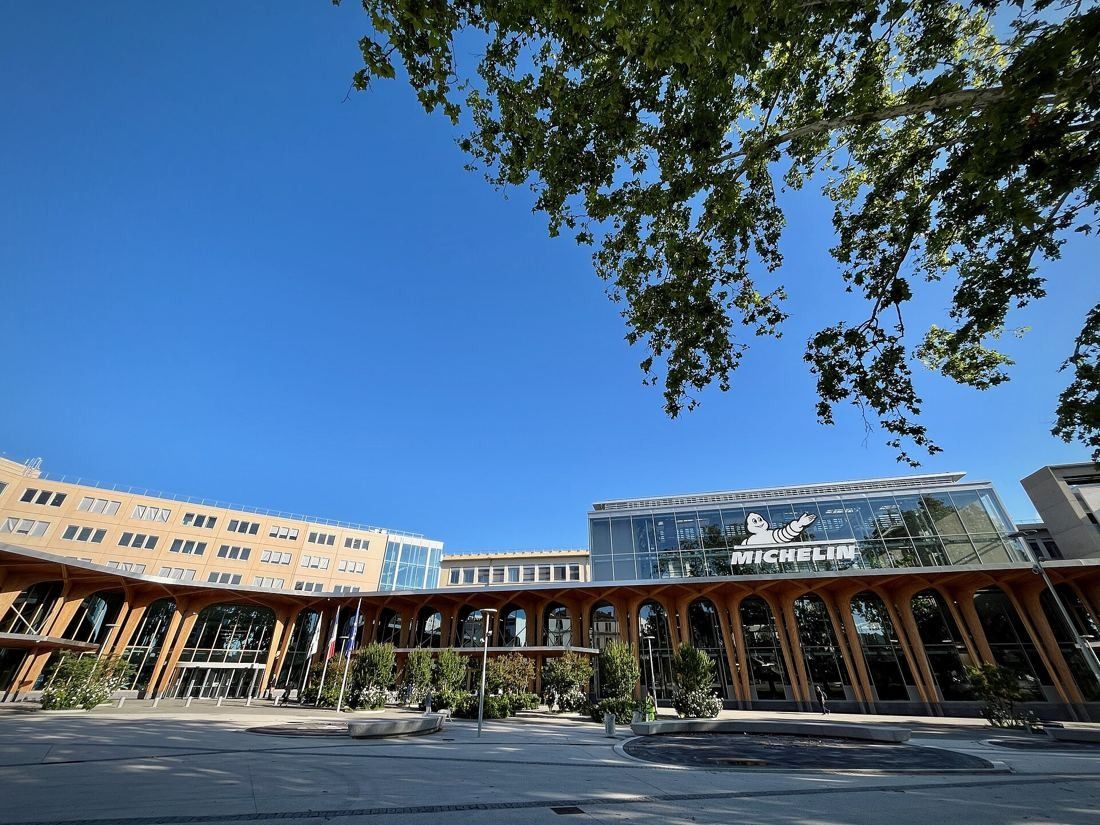 Michelin: Timber-arched welcome building and older office blocks under a blue sky, framed by tree branches