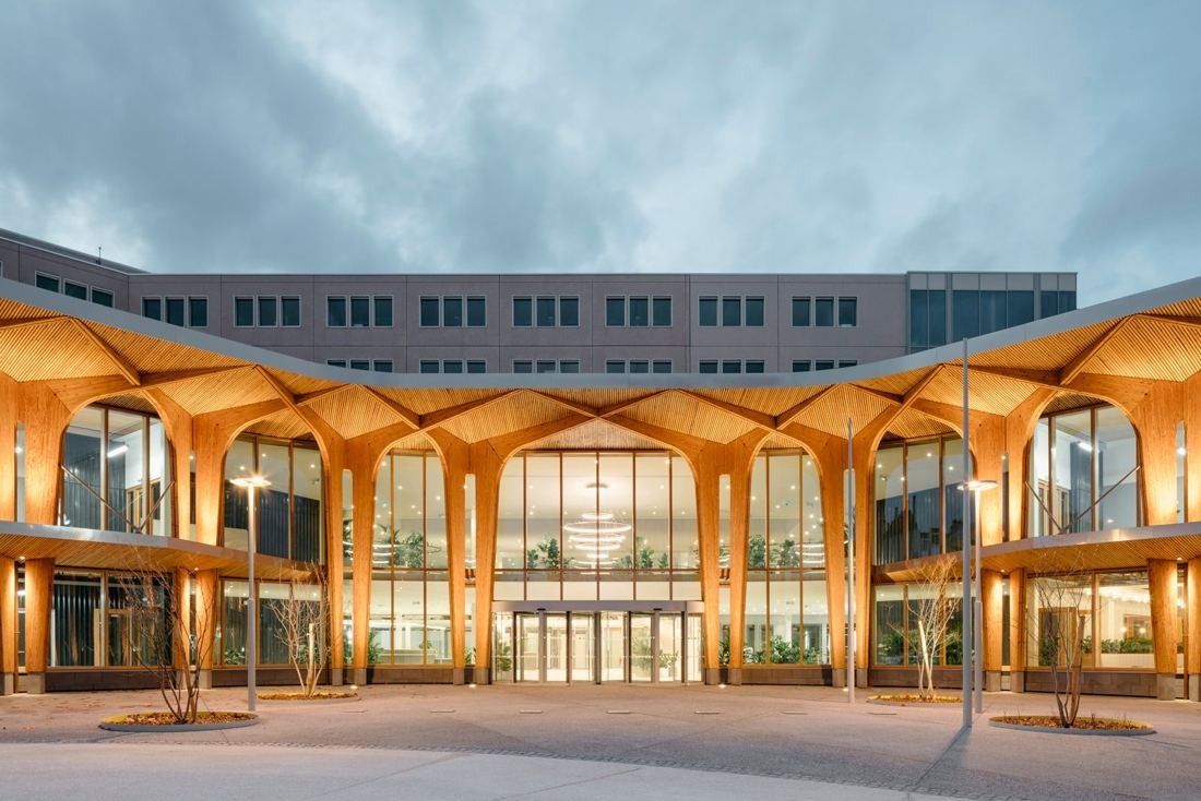 Two-storey timber façade with branching wooden arches, lit from inside at dusk, in front of an older office block