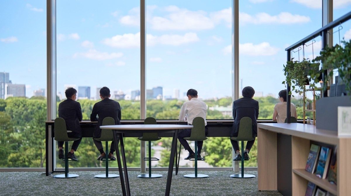Mitsubishi Corporation: People seen from behind at high tables facing a wall of windows over green trees