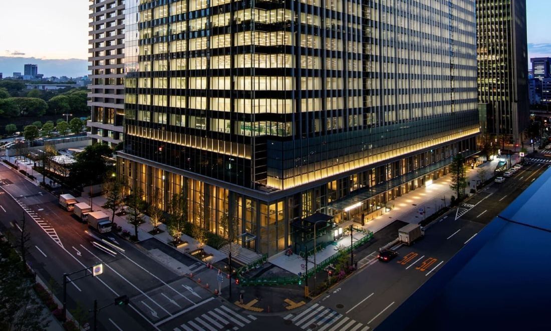 Stone-and-glass office tower lit at dusk beside a wide avenue and the palace moat