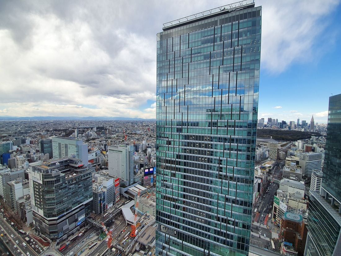MIXI: Glass skyscraper rising above low-rise Shibuya under a cloudy sky