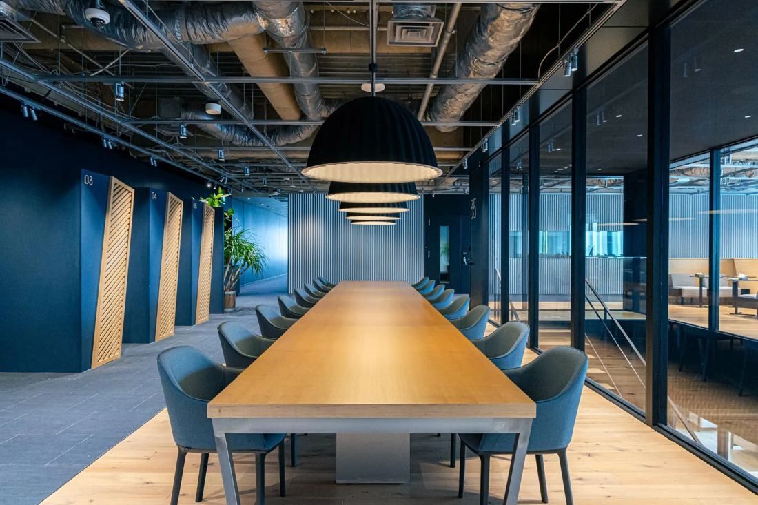 MIXI: Long wooden meeting table under a large pendant lamp beside glass rooms