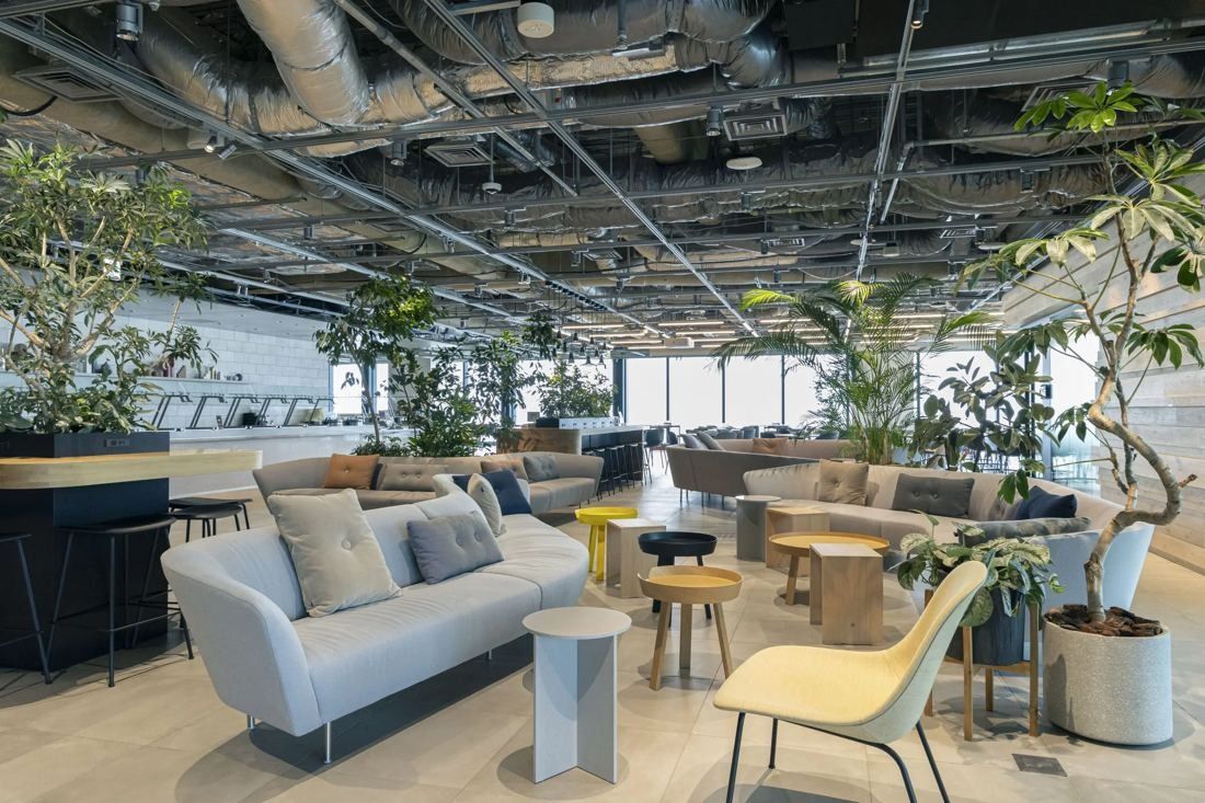 MIXI: Lounge with sofas, small tables and many plants under an exposed ceiling