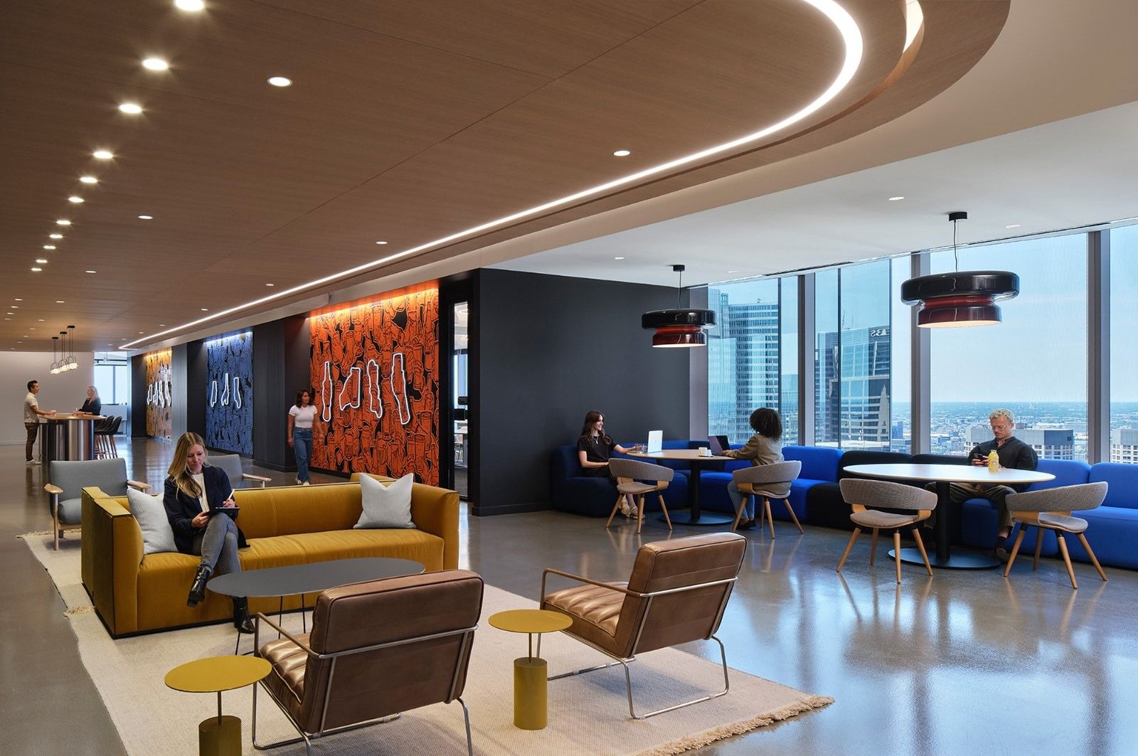 A long lounge with mustard and blue sofas beside tall windows and a red brand wall