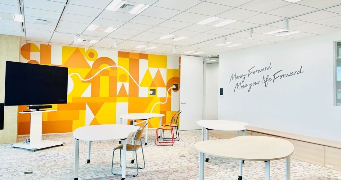 Money Forward: Bright meeting space with round white tables, a screen and a wall painted with orange and yellow shapes