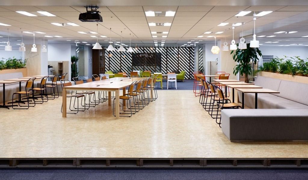 Large open room with long wooden tables, chairs and a light floor under rows of ceiling lights