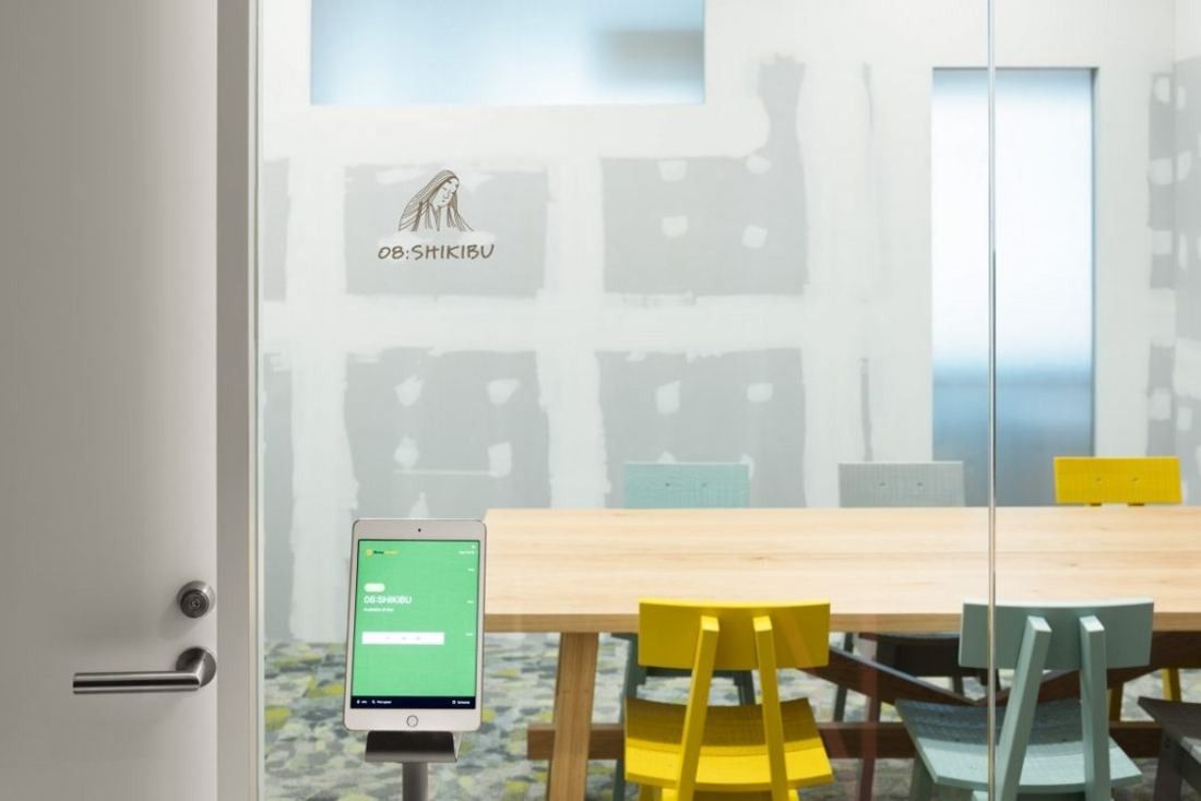 Money Forward: Meeting room with a light wooden table, yellow chairs and a tablet by the door