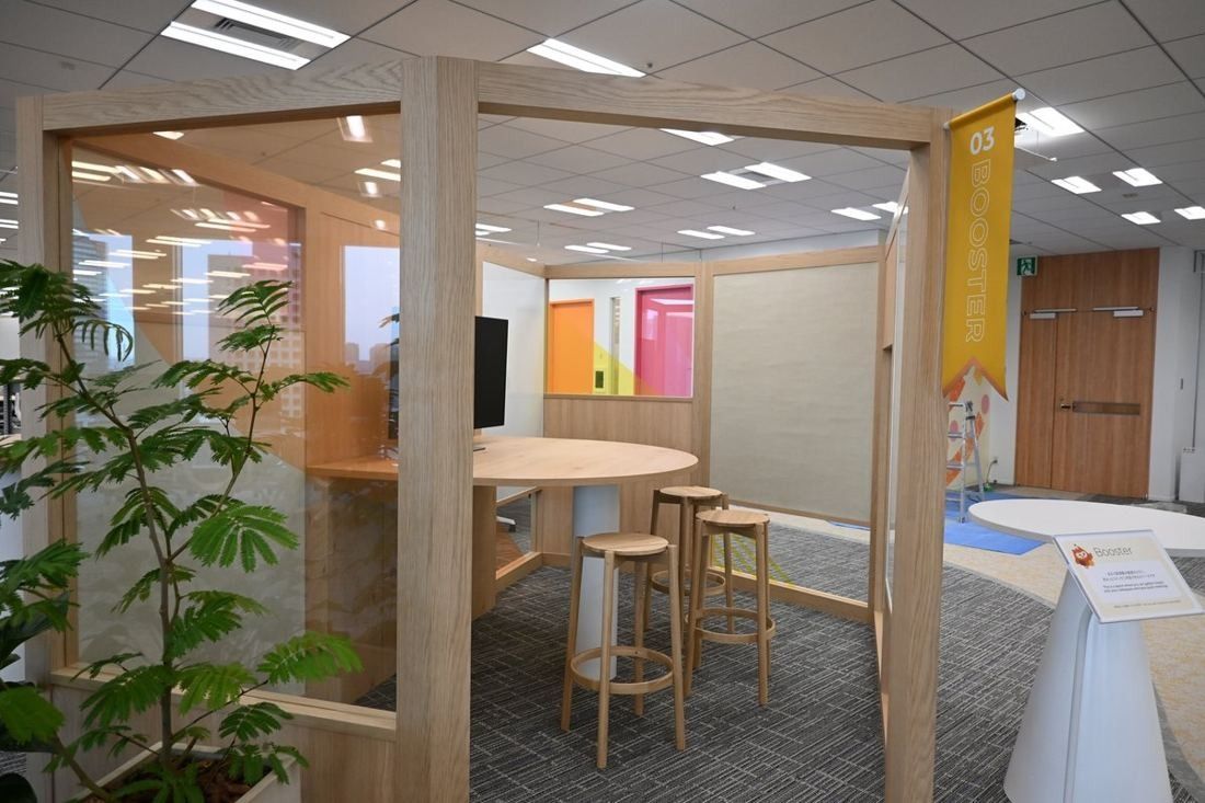 Money Forward: Open wooden pod with a high round table and stools, plants and a banner beside it