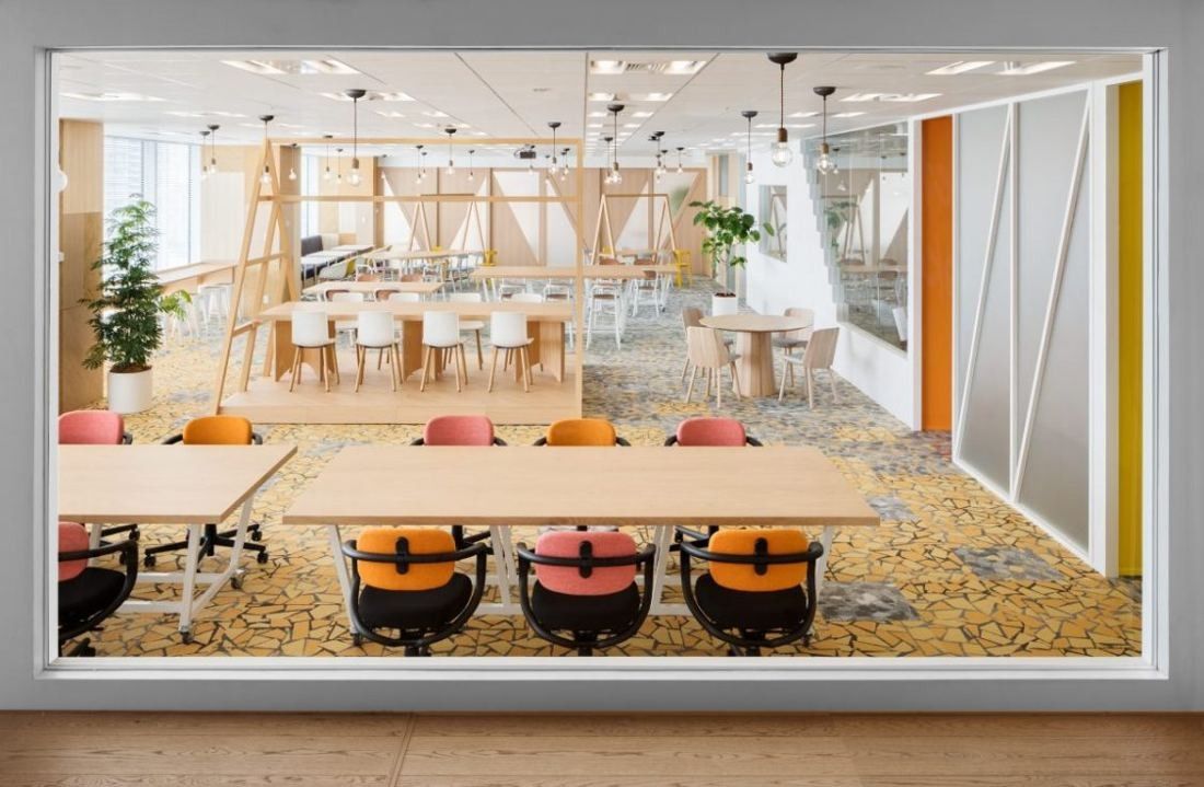 Money Forward: Open workspace with wooden tables and orange chairs seen through a framed glass opening