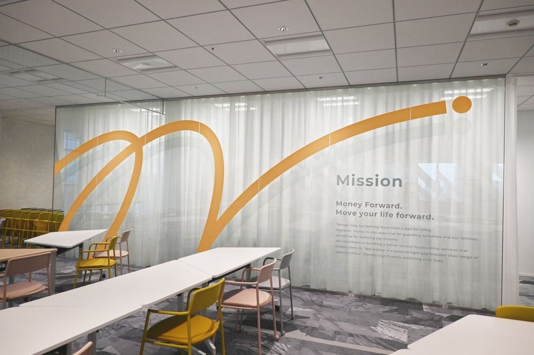 Money Forward: Room of long tables with yellow chairs and a sheer curtain printed with a gold line and text