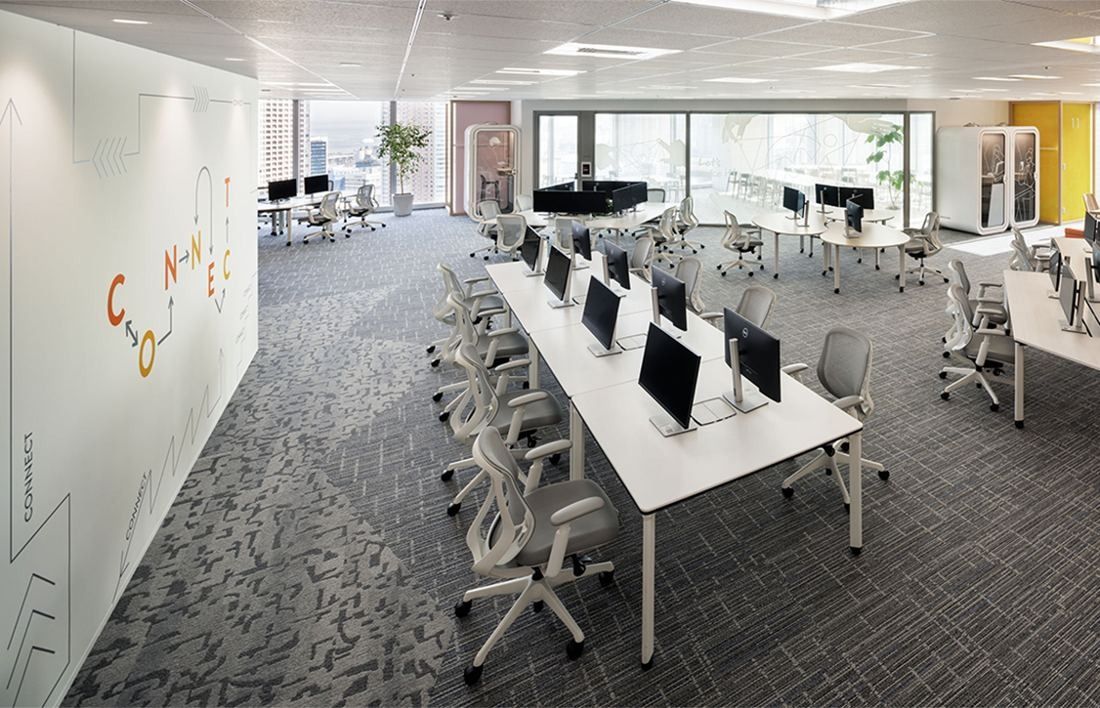 Money Forward: Rows of white desks with monitors on patterned grey carpet beside a white wall with lettering