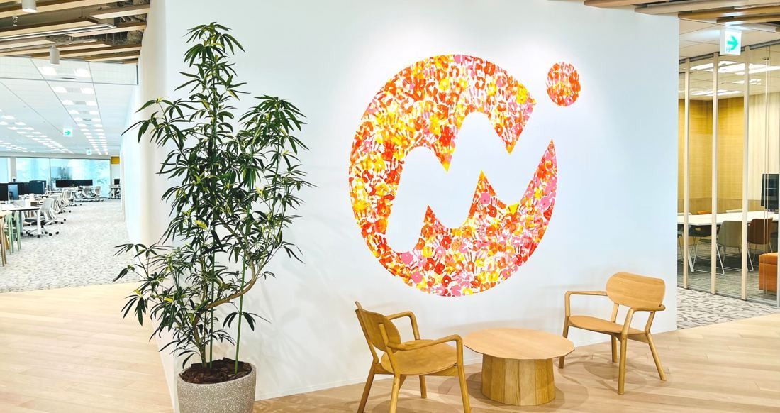 Money Forward: White wall with a flower-pattern logo, a potted plant and two wooden lounge chairs