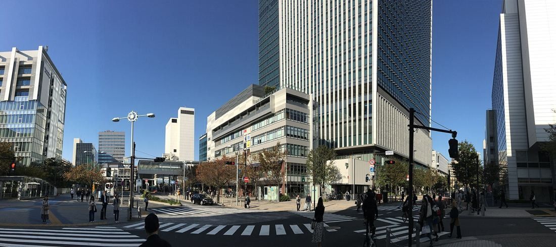 Money Forward: Wide street crossing in front of a station with high-rise towers on the right under a blue sky