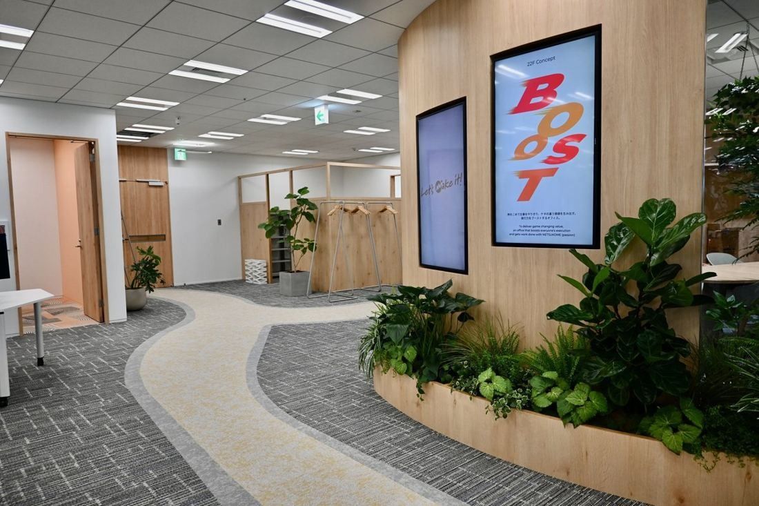 Money Forward: Winding path through grey carpet past a planter, a wooden column with a digital poster and meeting-room doors