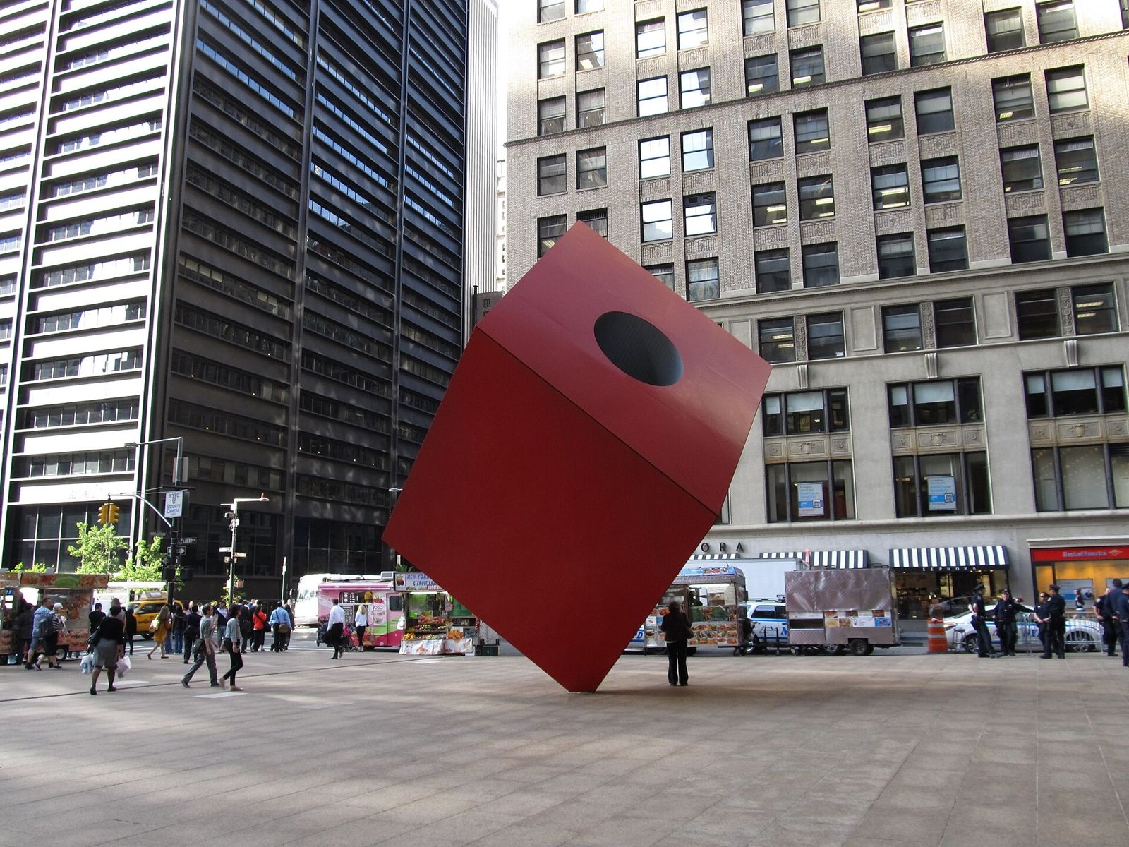 Red cube sculpture in front of a dark office tower on Broadway