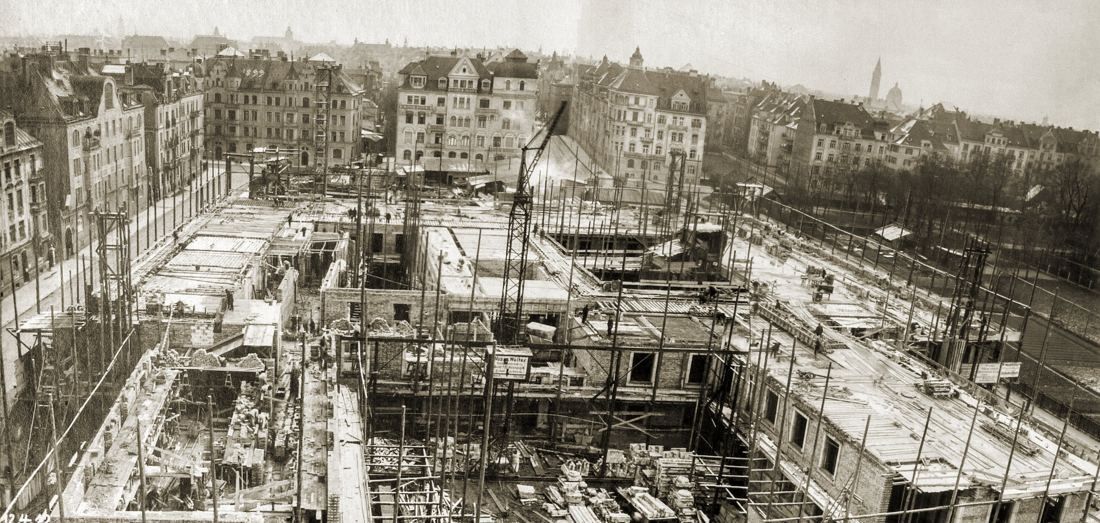 Wide black-and-white view over a large building site with scaffolding and a crane