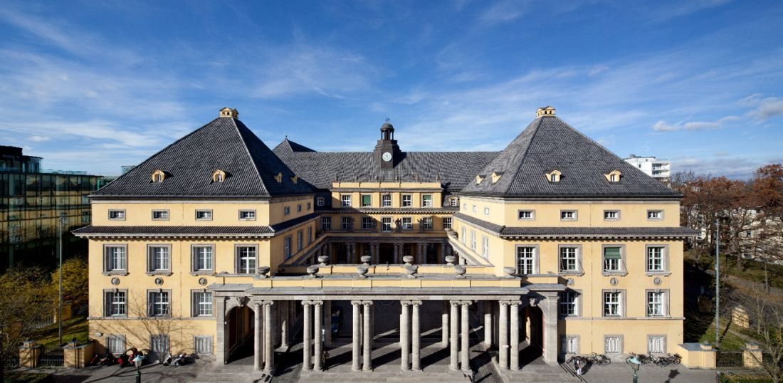 Yellow historicist building with two steep pyramid roofs, a clock turret and a columned entrance, seen from above