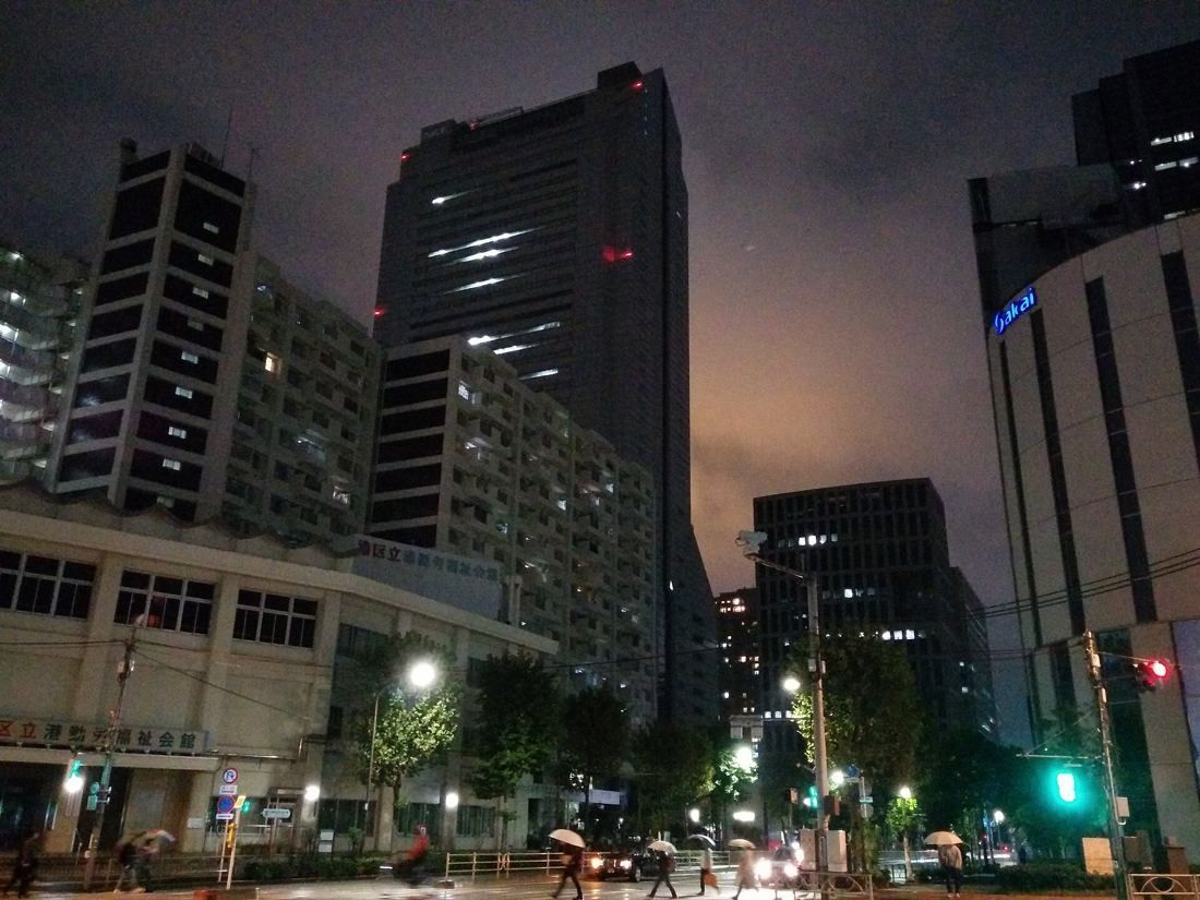 NEC: Dark tower behind apartment blocks at a rainy night crossing with umbrellas