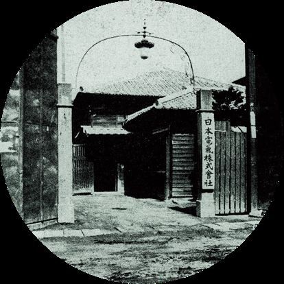 Old black-and-white photo of a gate with a lamp and a vertical company signboard