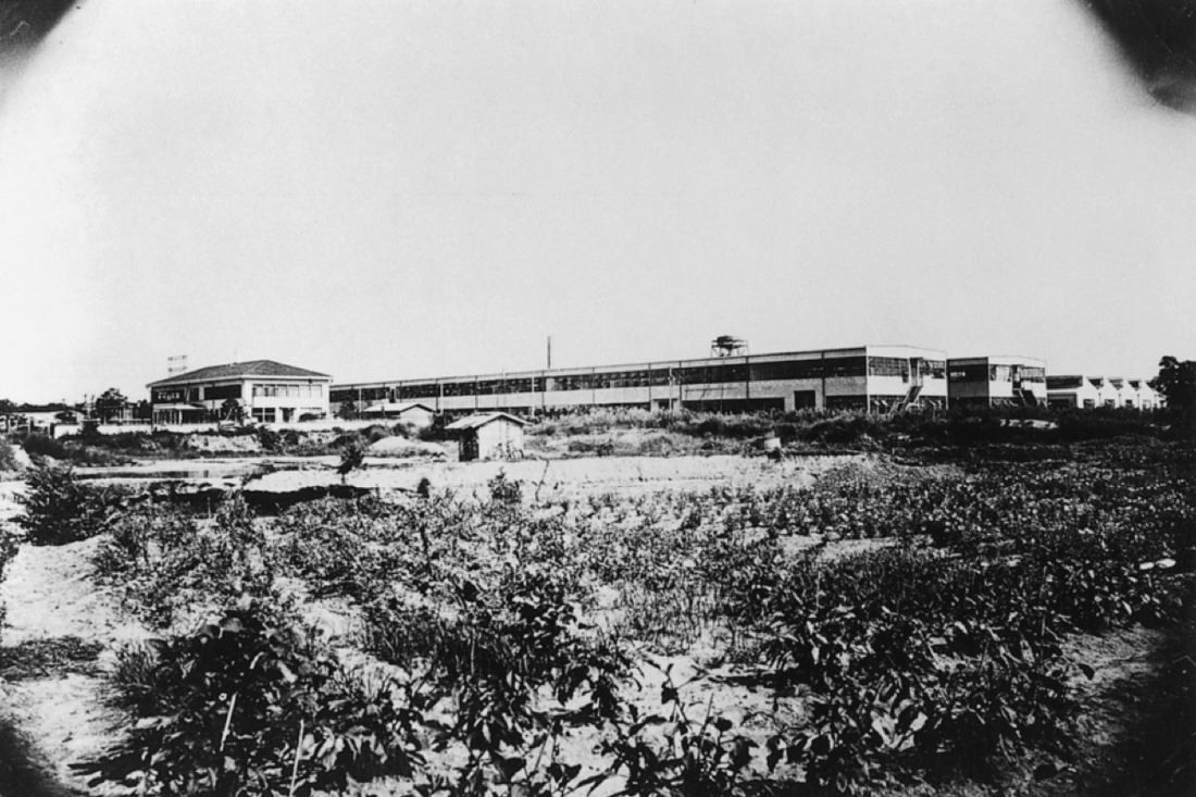 Old black-and-white photo of low factory buildings beyond open fields
