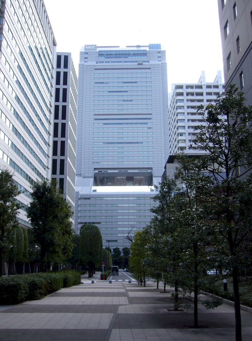 NEC: Tall pale tower with a large opening near its base, seen down a tree-lined plaza