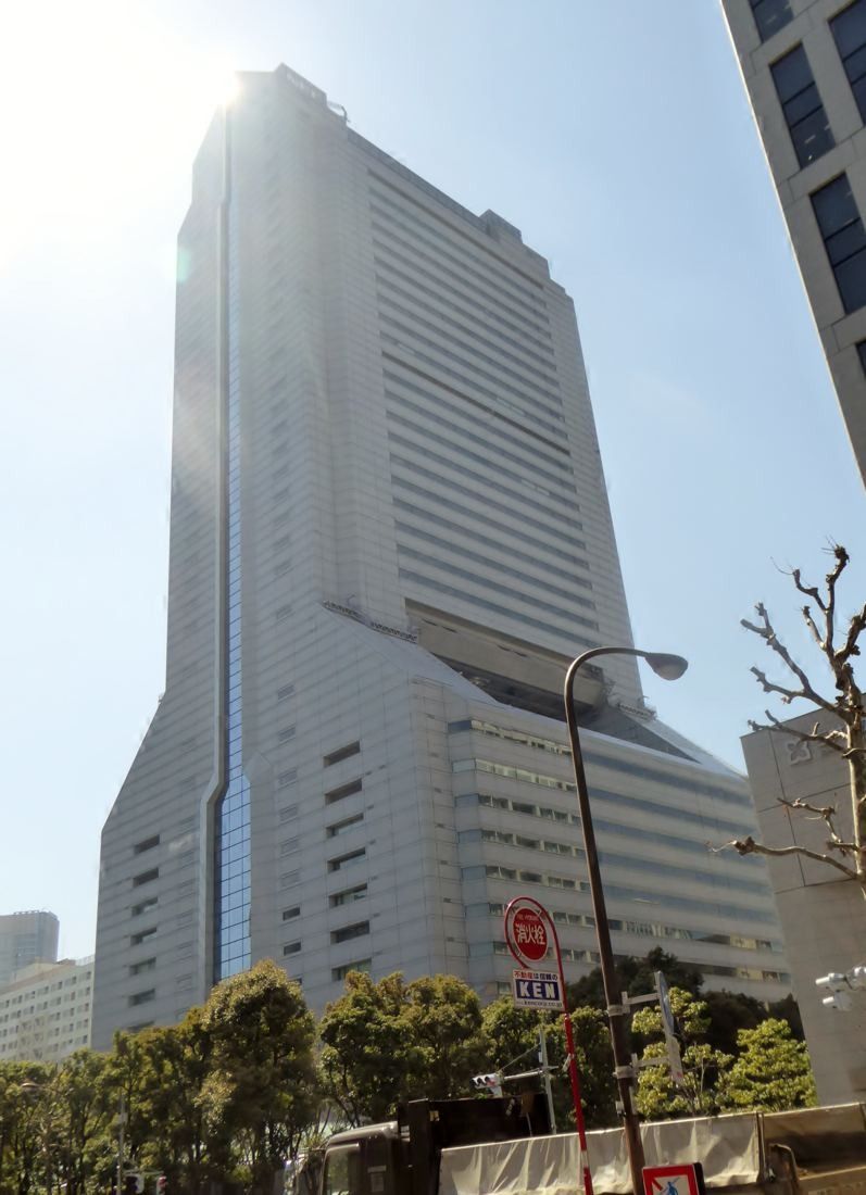 NEC: Tapering tower rising above trees and a street, seen from below
