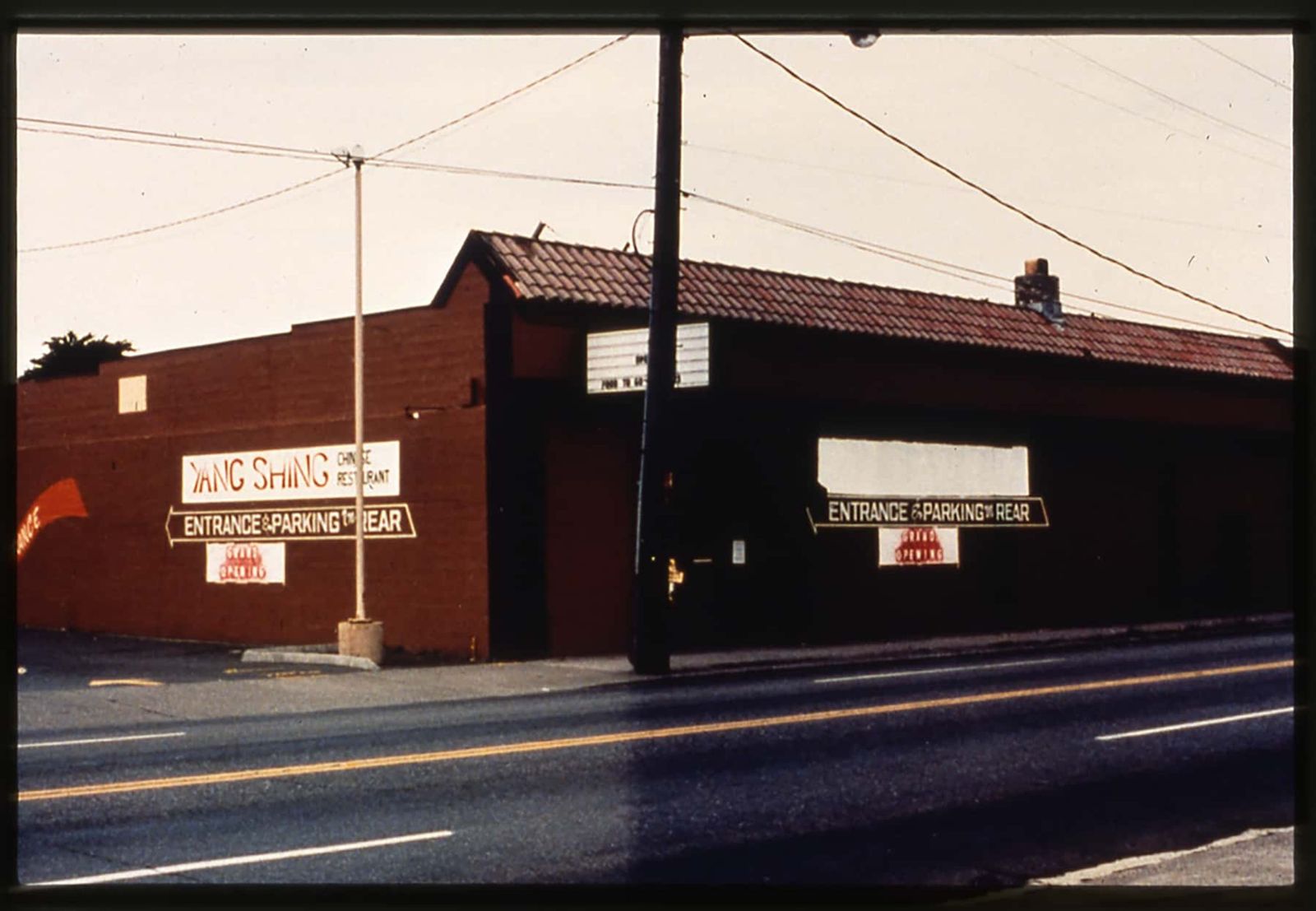 Low brick building with a tiled roof on a street corner, with 'Yang Shing' restaurant and 'Entrance & Parking in Rear' signs, power lines overhead
