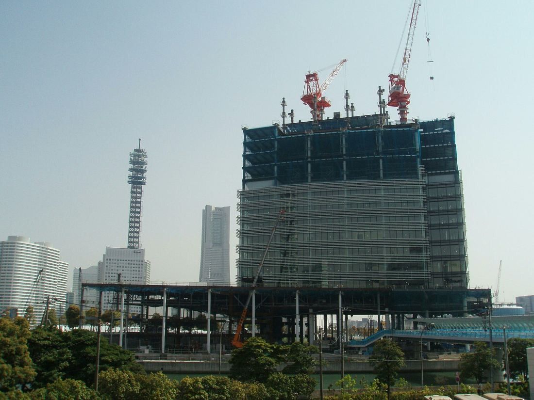 Steel frame of a high-rise with two red cranes on top, a tower visible in the distance