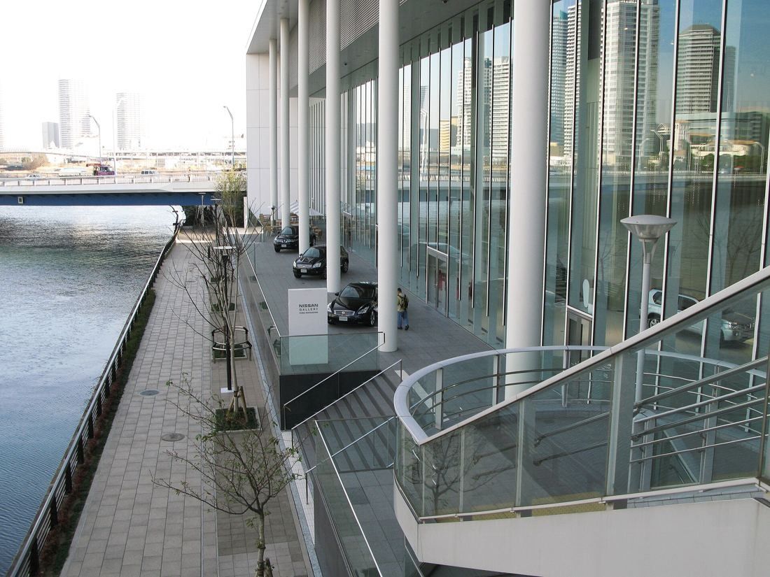 Nissan: Tall glass wall and white columns along a canal-side walkway, with cars parked under the overhang