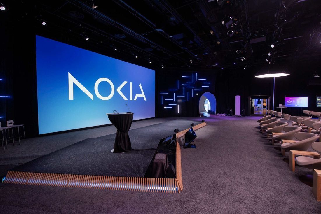 Nokia: Dark presentation hall with a large screen showing the Nokia logo, rows of lounge seats and low wooden benches