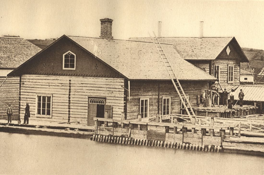 Old photograph of wooden mill buildings on a log quay by the water, with a ladder on the roof