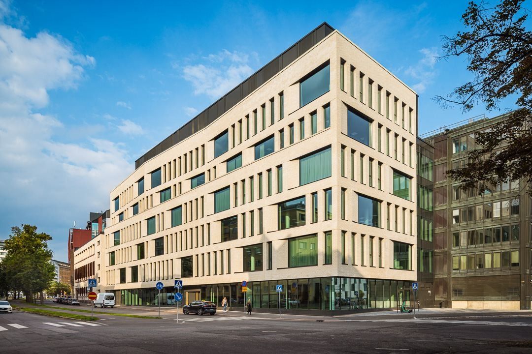 Eight-storey office building with a pale brick facade and rows of tall windows on a street corner under blue sky