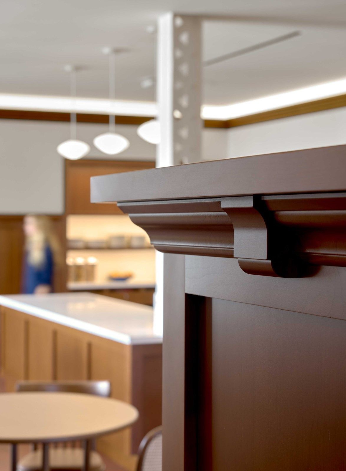 Notion: Detail of a carved wooden counter cap with a riveted column and pendant lights behind