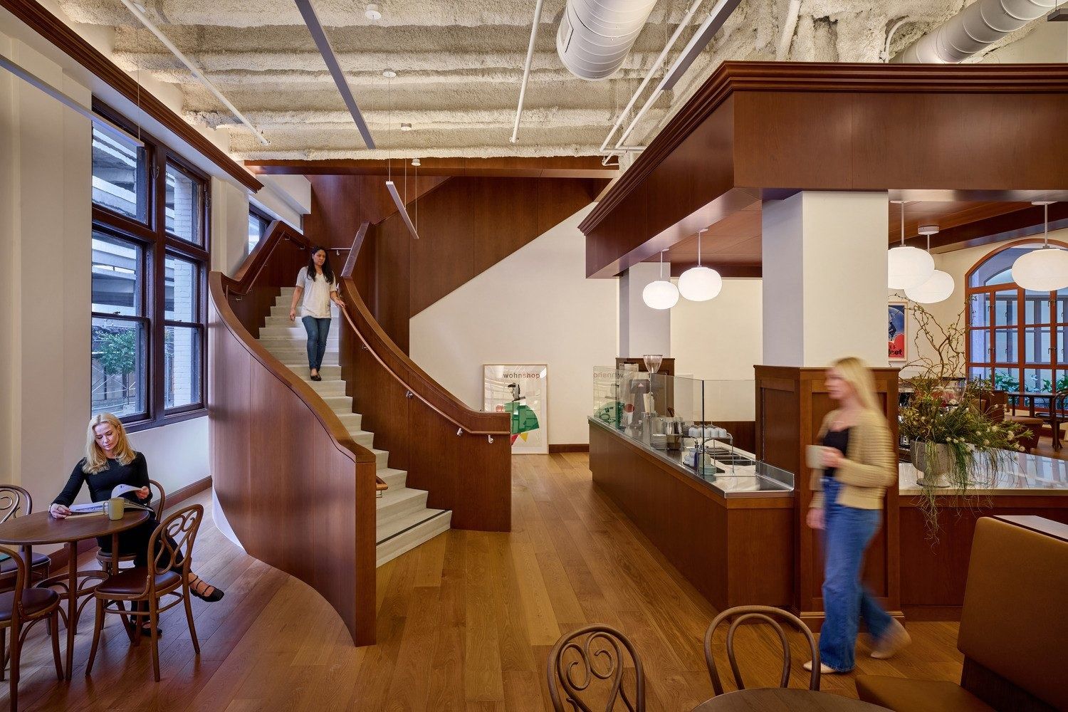Double-height space with a curved wooden staircase beside a coffee bar, globe pendant lights and café tables