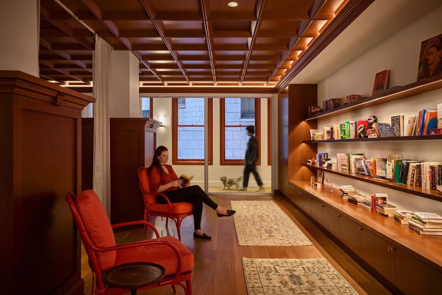 Notion: Narrow room with a coffered wood ceiling, long lit bookshelves, red armchairs and patterned runners