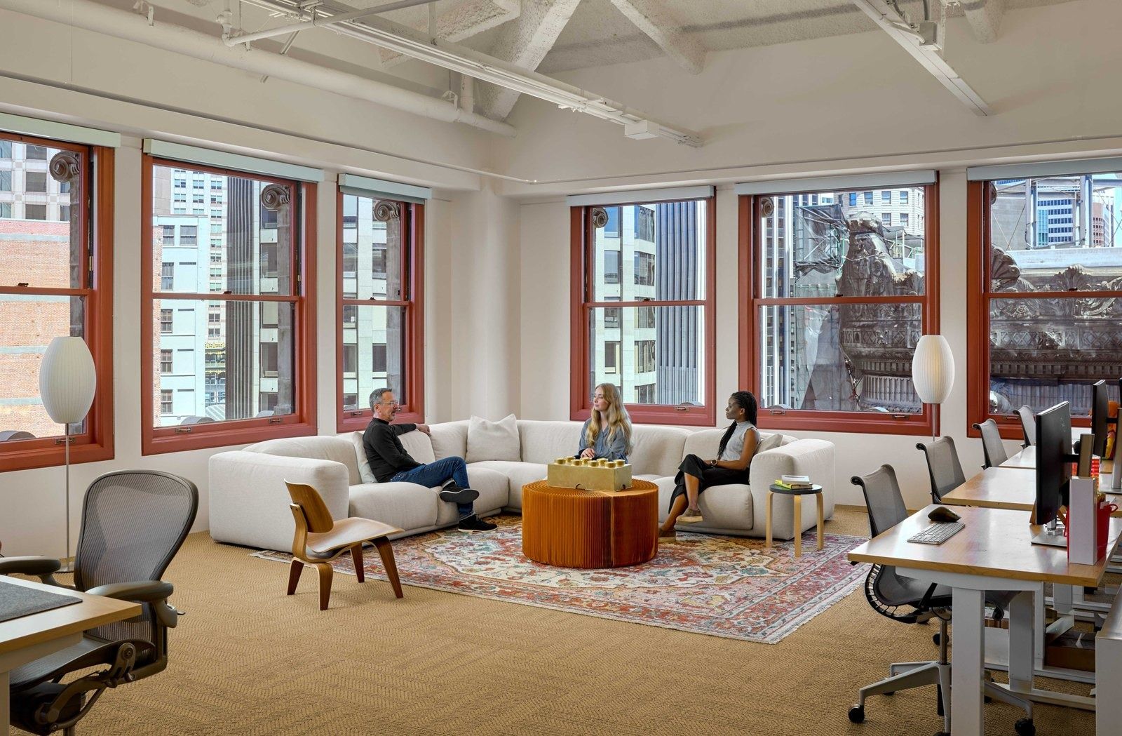 Notion: Open floor with a white sectional sofa on a rug, desks with monitors and tall red-framed windows onto the city