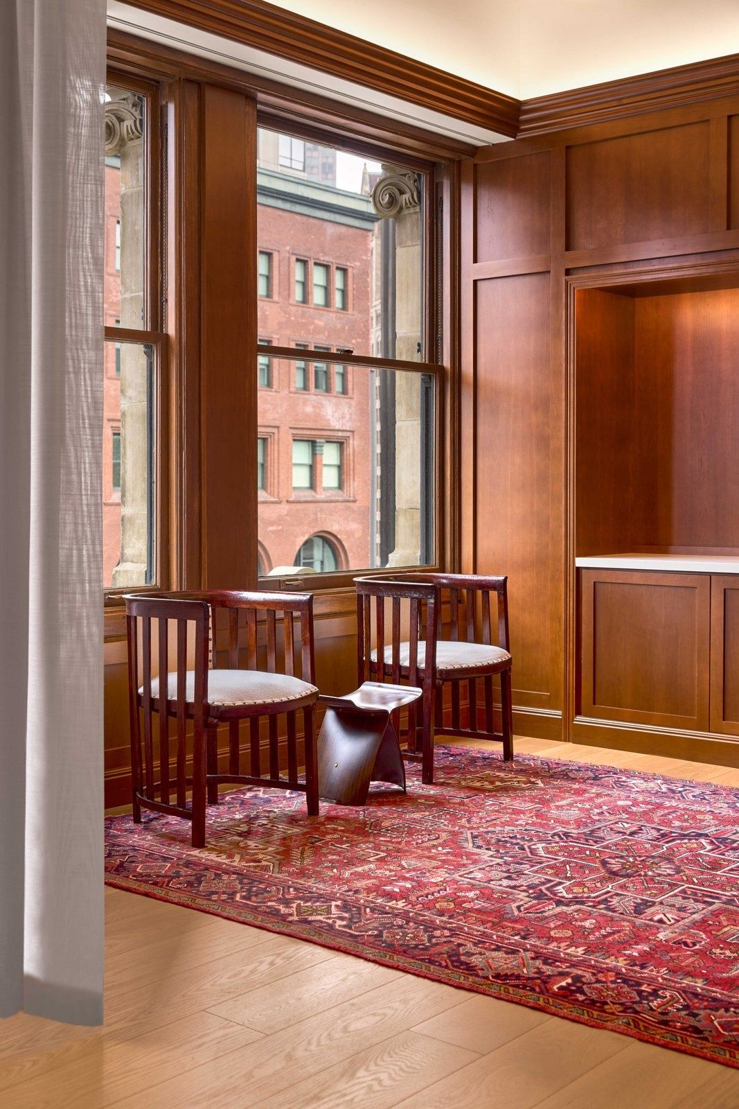 Notion: Window corner with two wooden armchairs on a red rug, wood paneling and a view of brick buildings
