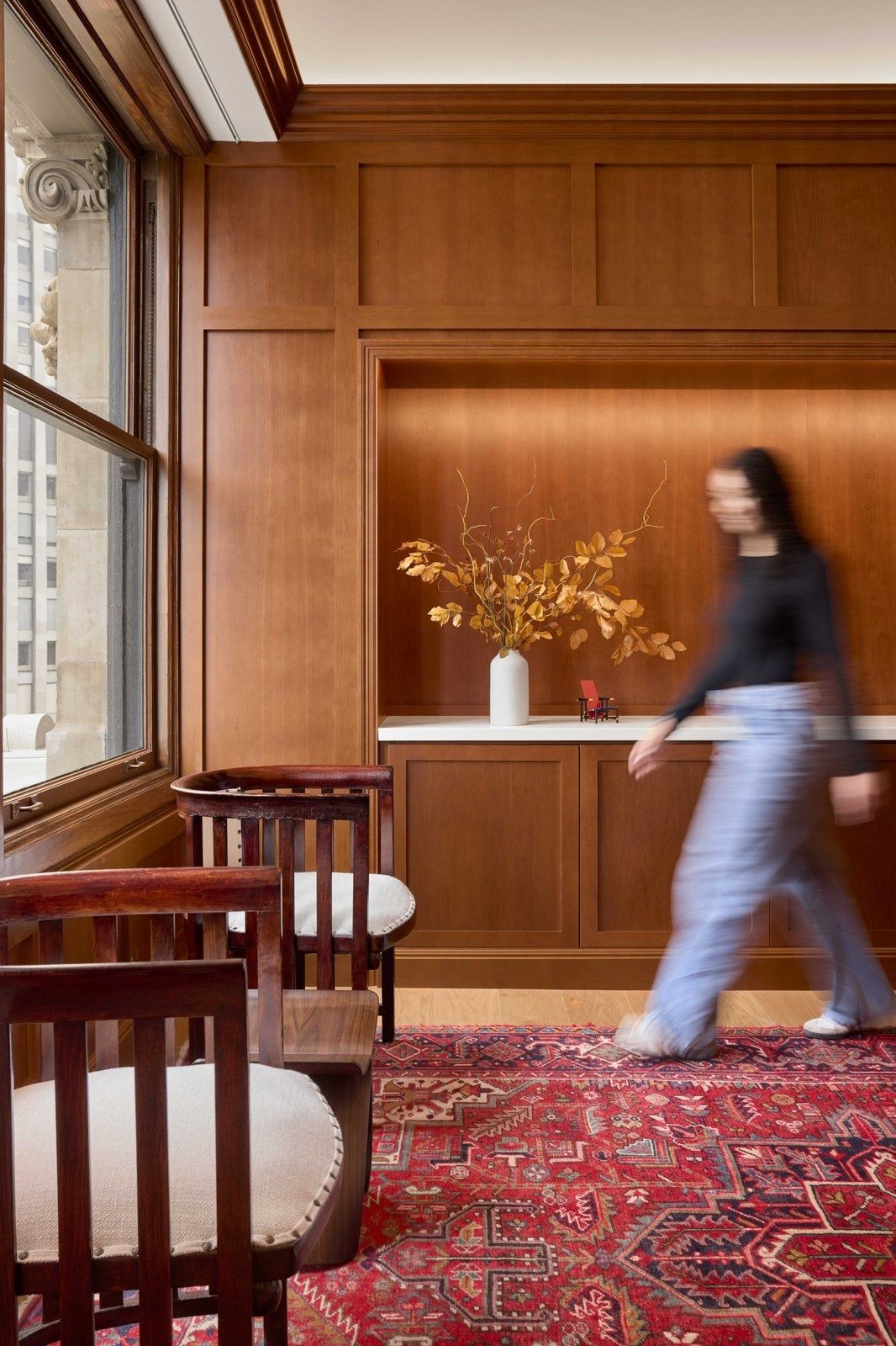Notion: Wood-paneled wall with a lit niche and a vase of branches, wooden chairs on a red rug