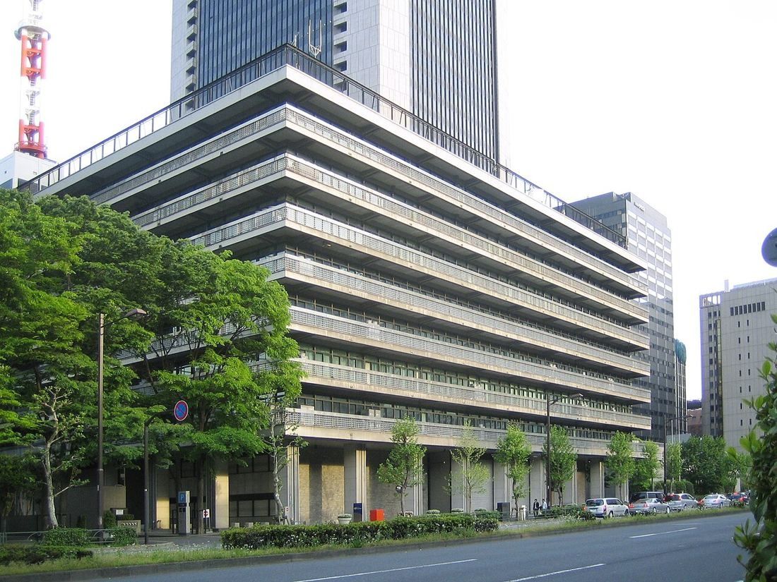 Nine-storey office building with deep stacked balconies beside a tree-lined avenue