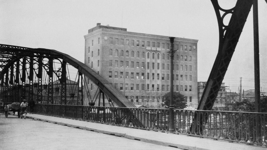 Black-and-white photo of a seven-storey brick office building seen from a steel arch bridge