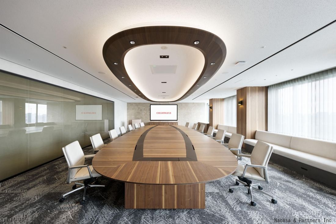 Okamura: Long oval wooden boardroom table with white chairs under an oval ceiling light